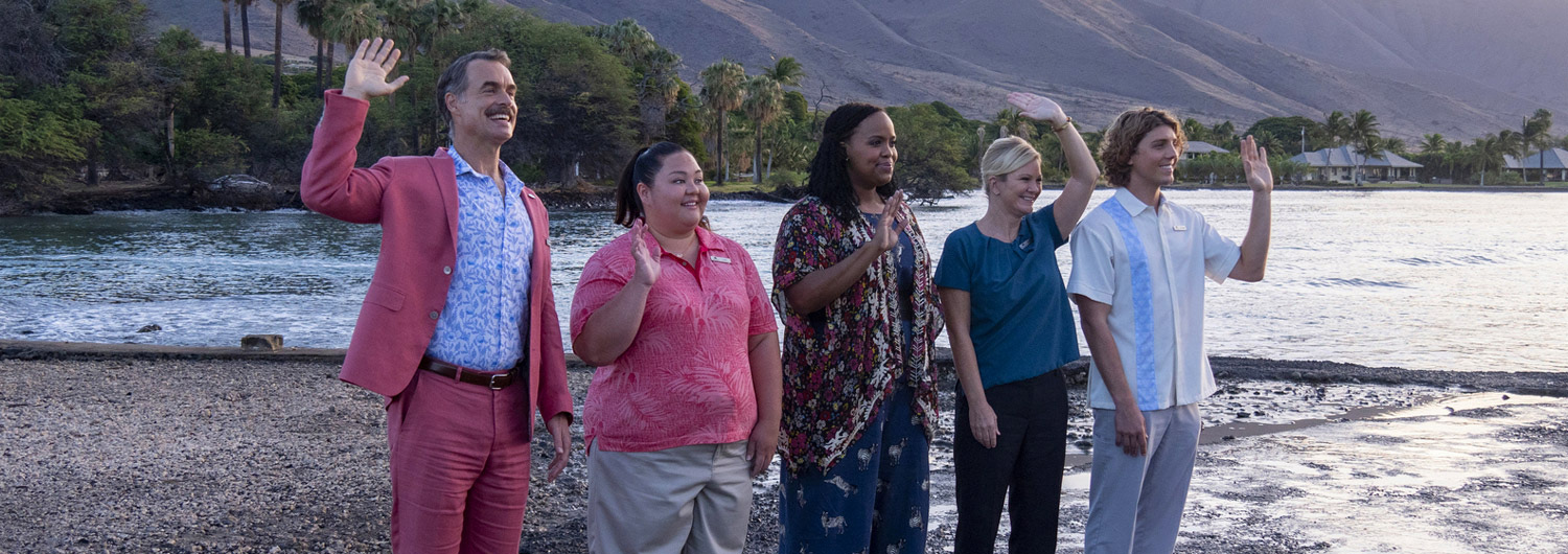 Cast members of The White Lotus waving their hands with water and a Hawaiian landscape behind them